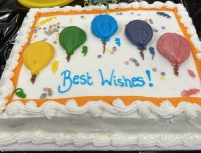 Picture shows a white cake decorated with colourful balloons and 'Best Wishes'.